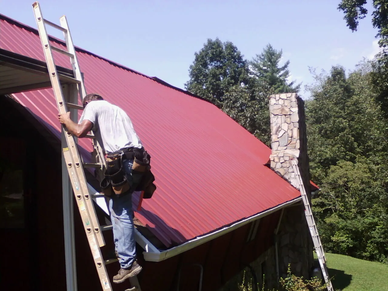 Roofer on a ladder beside a red metal roof and stone chimney — Soffit & Fascia Repair by Roofing Brothers in East Granby